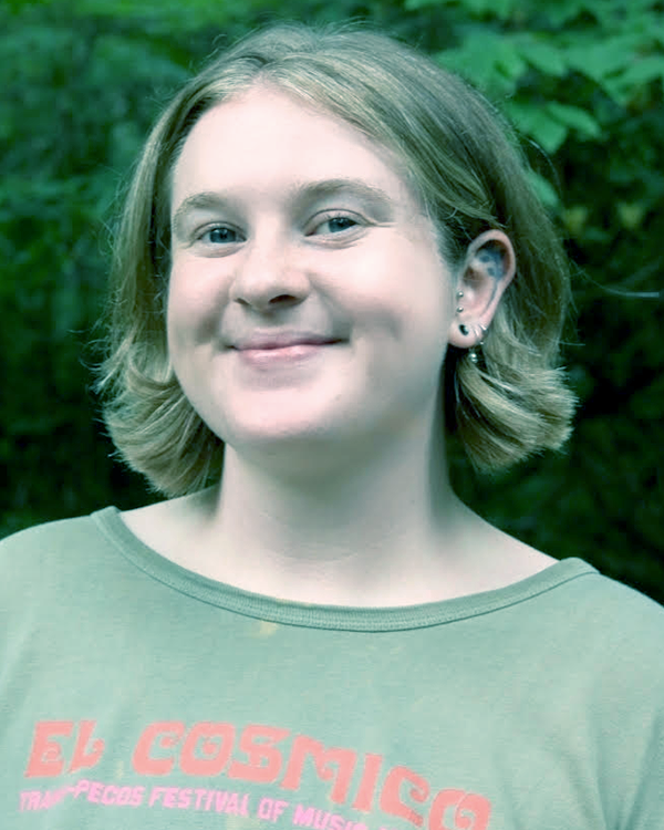 A smiling young woman with short blonde hair, wearing earrings and a green T-shirt with pink text, standing outdoors with green foliage in the background.