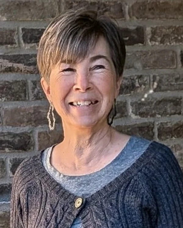 Smiling woman with short hair, earrings, and a gray sweater standing in front of a brick wall.