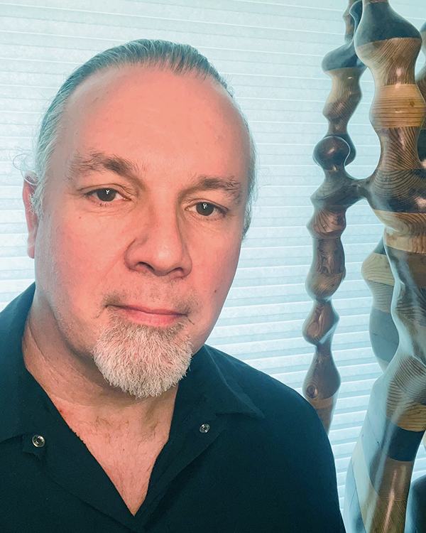 Close-up of a man with a goatee and long hair tied back, wearing a dark collared shirt, standing next to abstract wooden sculptures against a backdrop of window blinds.