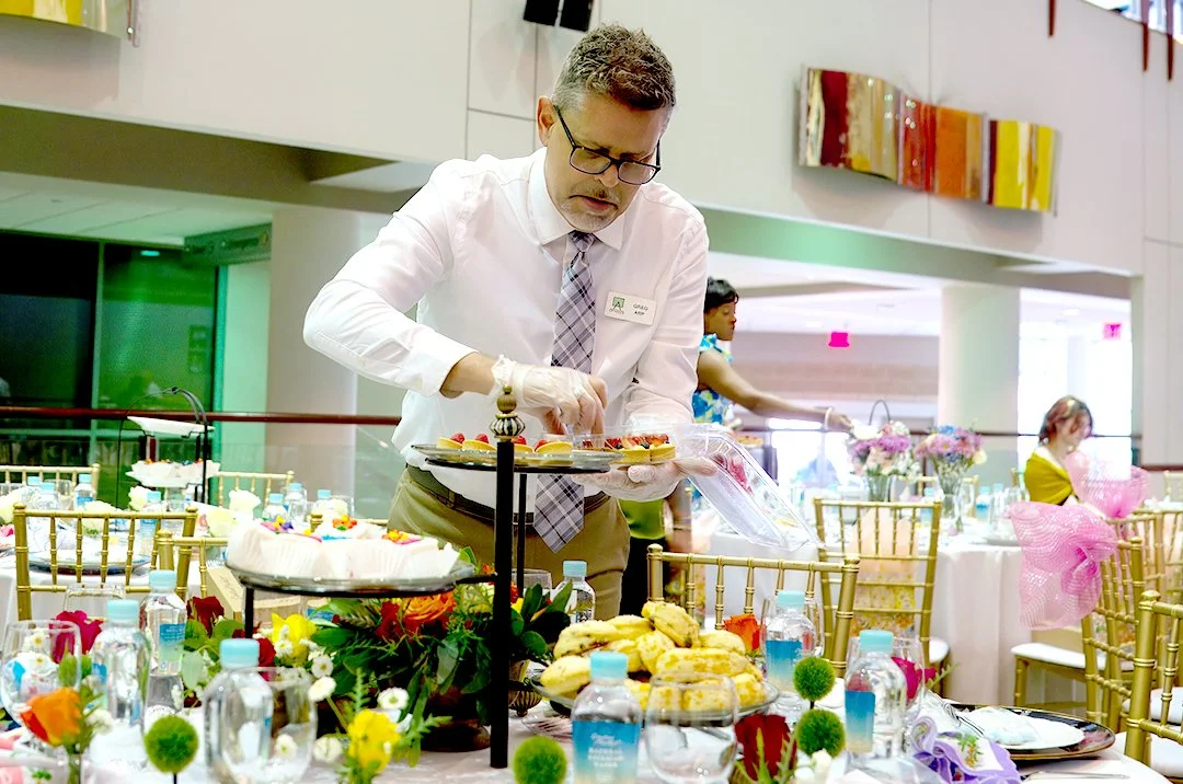 A man in a white shirt and tie setting up a dessert table at an event with floral arrangements and gold chairs.
