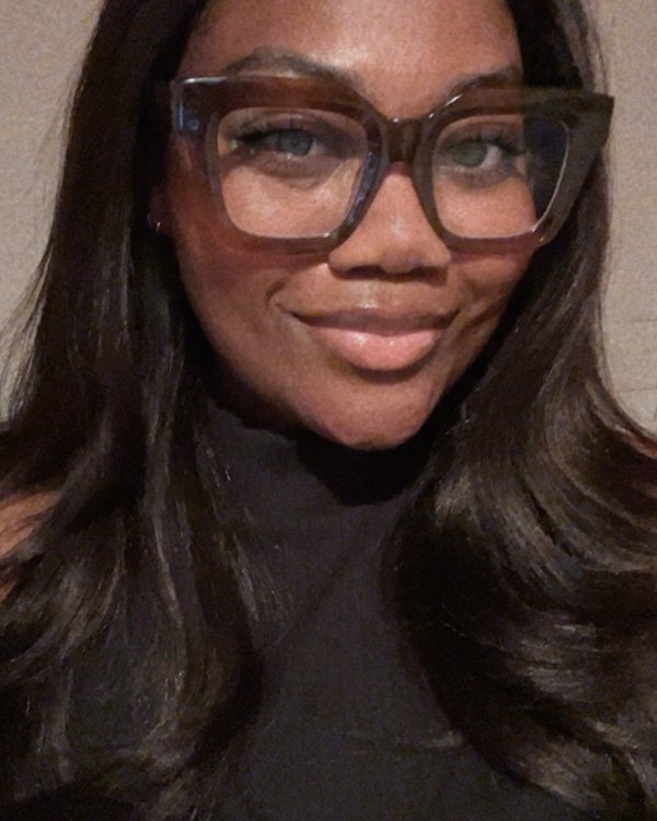 Close-up of a woman with long dark hair wearing large glasses and a black top, smiling at the camera.