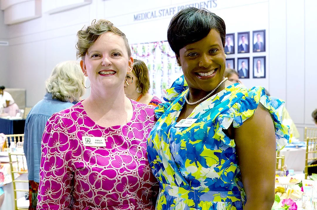 Two women smiling at an indoor event, both wearing colorful dresses. The woman on the left has a short curly hairstyle and the woman on the right has a short hairstyle. They are surrounded by other people and tables in a room with framed photographs 