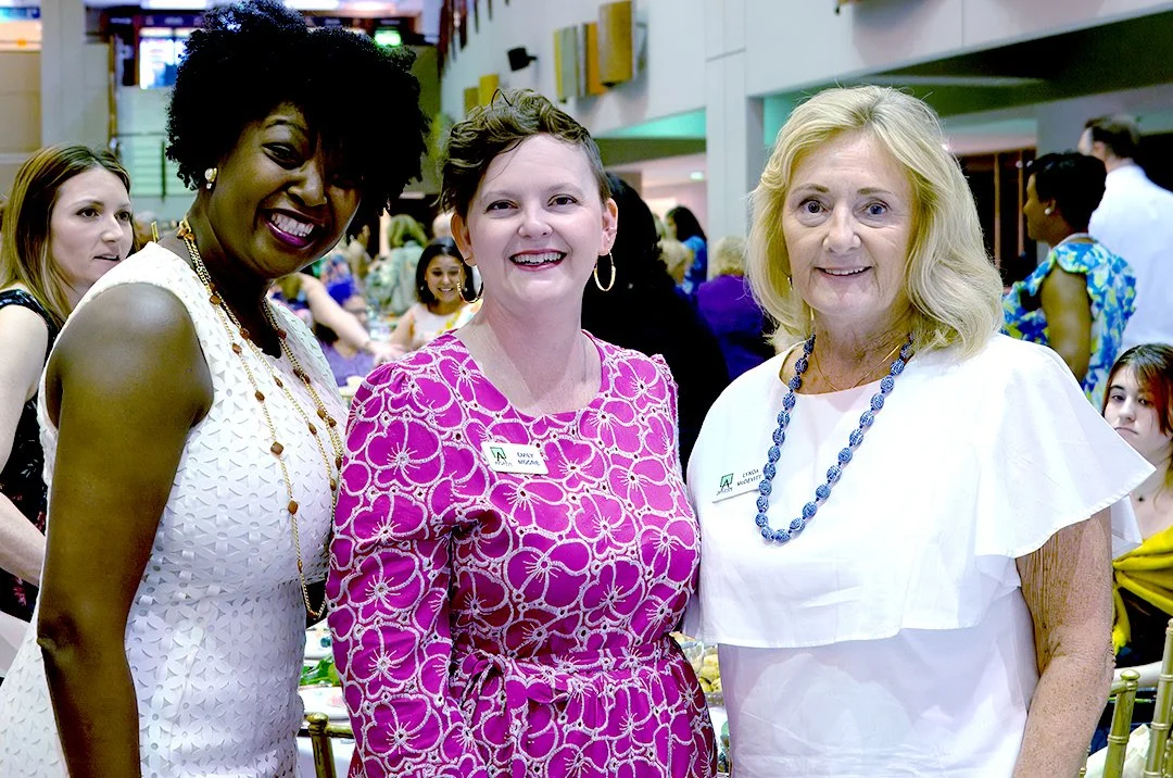 Three women smiling at a social event, surrounded by other attendees.