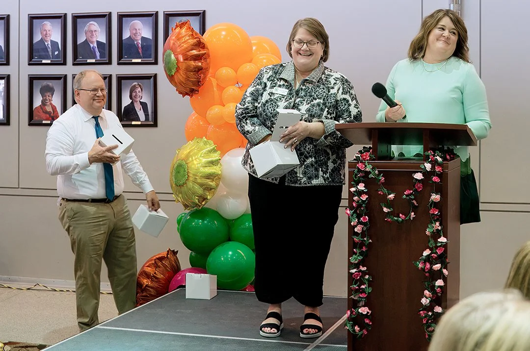Three people in an office setting, standing near a podium with floral decorations. They appear to be part of an event with colorful balloons and framed portraits on the wall. Each person is holding a white box, suggesting award or gift distribution.