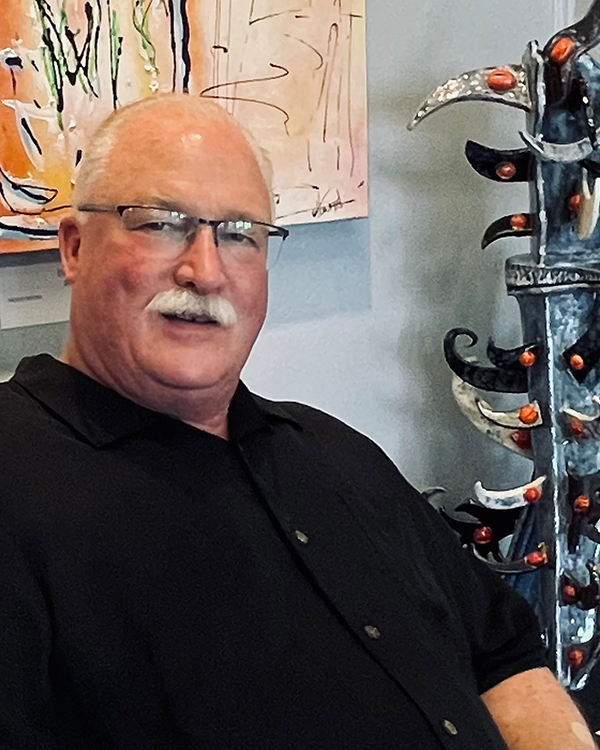 A man with glasses and a white mustache smiling, wearing a black shirt, seated near abstract artwork and a decorative metal sculpture with curved black and white elements and small orange accents.