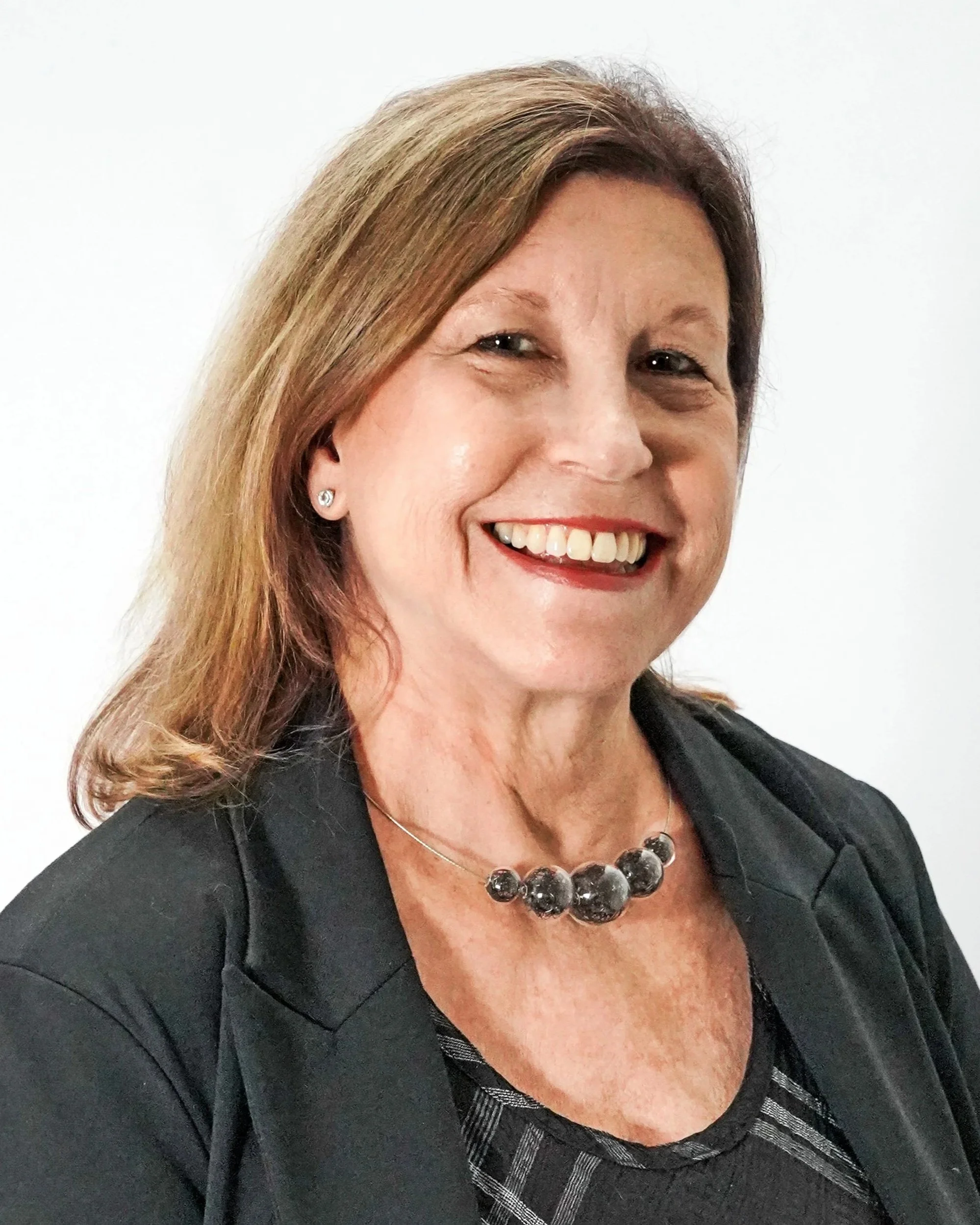 A smiling middle-aged woman with light brown hair, wearing a black blazer, a beaded necklace, and small earrings, against a plain white background.