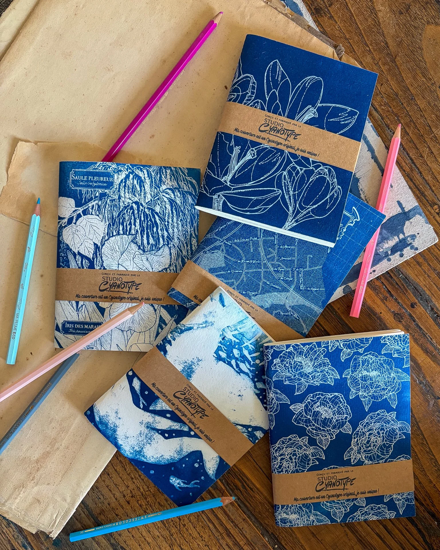 Carnets à dessins avec des illustrations en bleu et blanc et des emballages en papier kraft, disposés sur une surface en bois. Deux stylos colorés, un bleu et un rose, sont placés à côté des carnets.