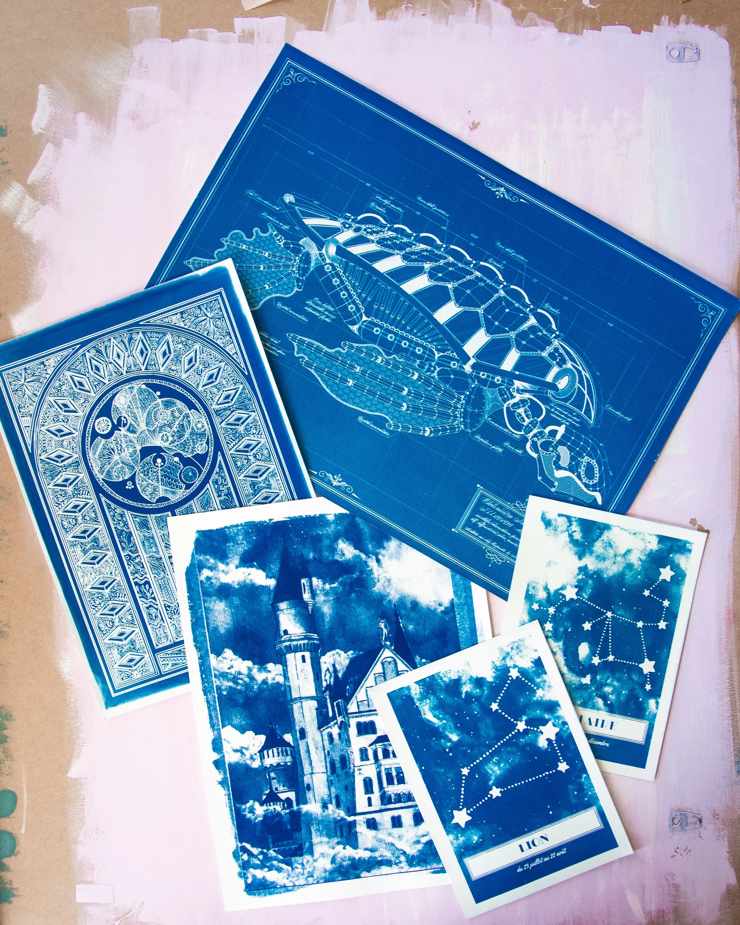 Une collection de cartes de tirages en cyanotype Bleu de Prusse disposées sur une surface peinte en rose pâle, avec des détails art déco et des illustrations de paysages et symboles.