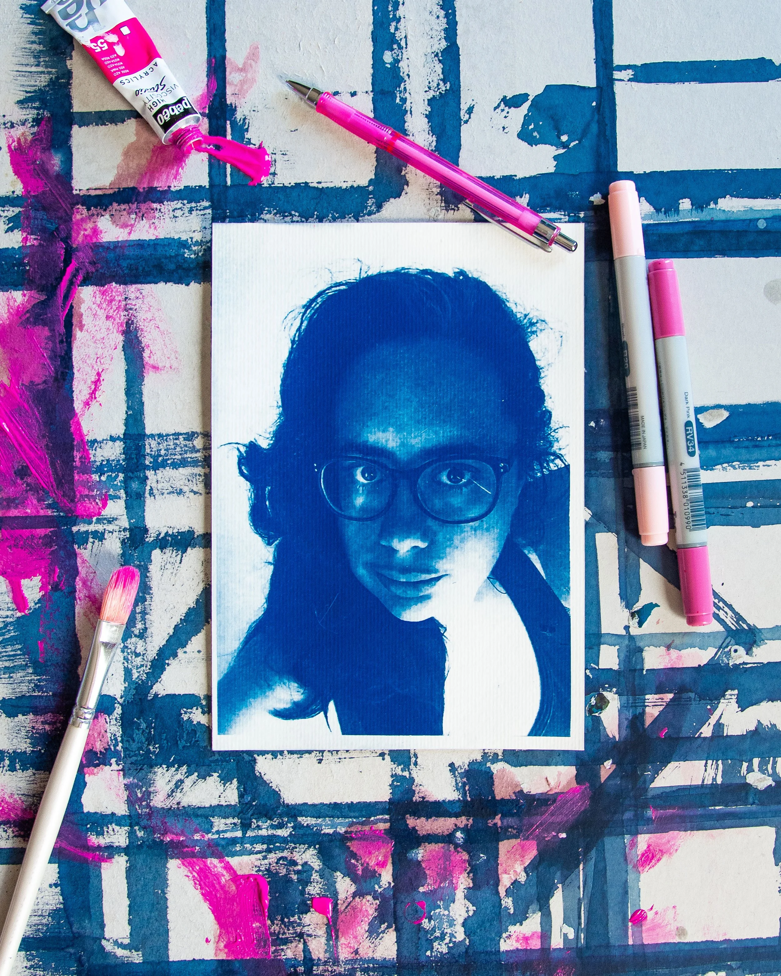 Une photo d'un tirage cyanotype bleu de prusse posé sur un carton peint avec des traces roses et blanches, incluant un portrait stylisé d'une personne portant des lunettes