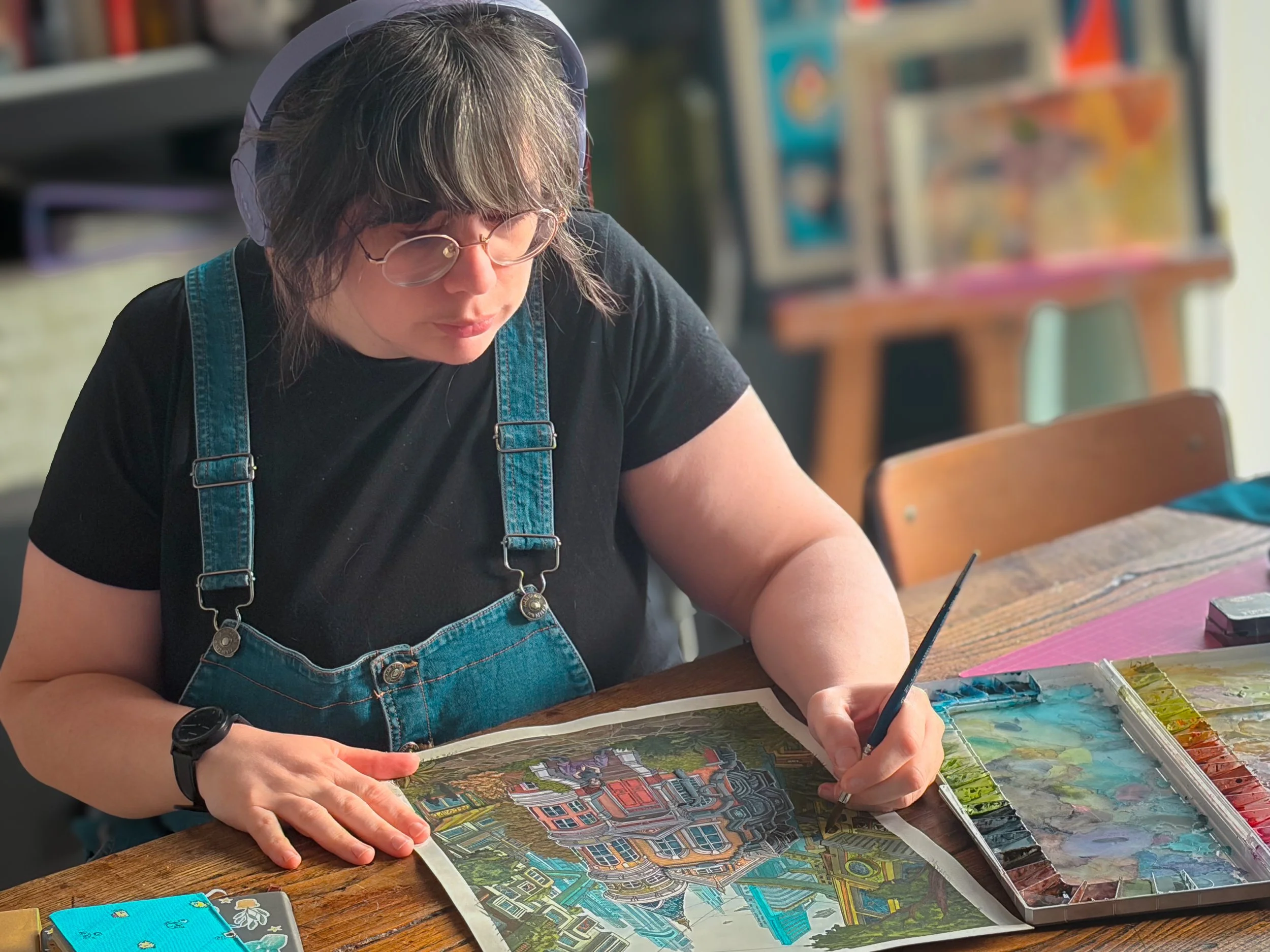 Une femme avec des lunettes, des écouteurs et une montre, portant un jean et un t-shirt noir, est en train de peindre une toile représentant une scène urbaine colorée dans un atelier d'artiste avec des livres en arrière-plan.