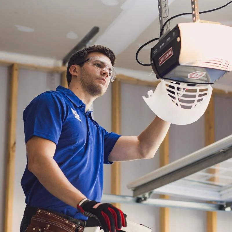 Technician in blue shirt installing or repairing a garage door opener with safety goggles and tools.