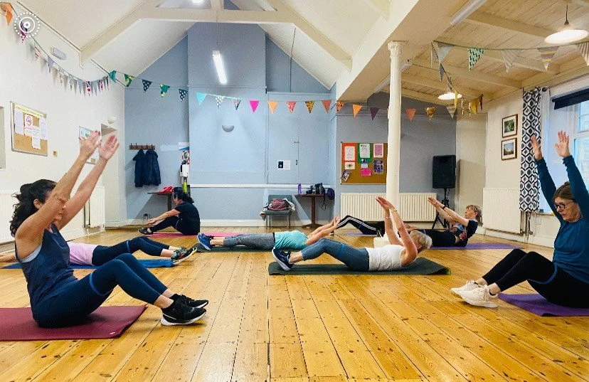 Women participating in a seated yoga or exercise class, sitting on mats, with arms raised overhead in a room decorated with bunting and wall decorations.