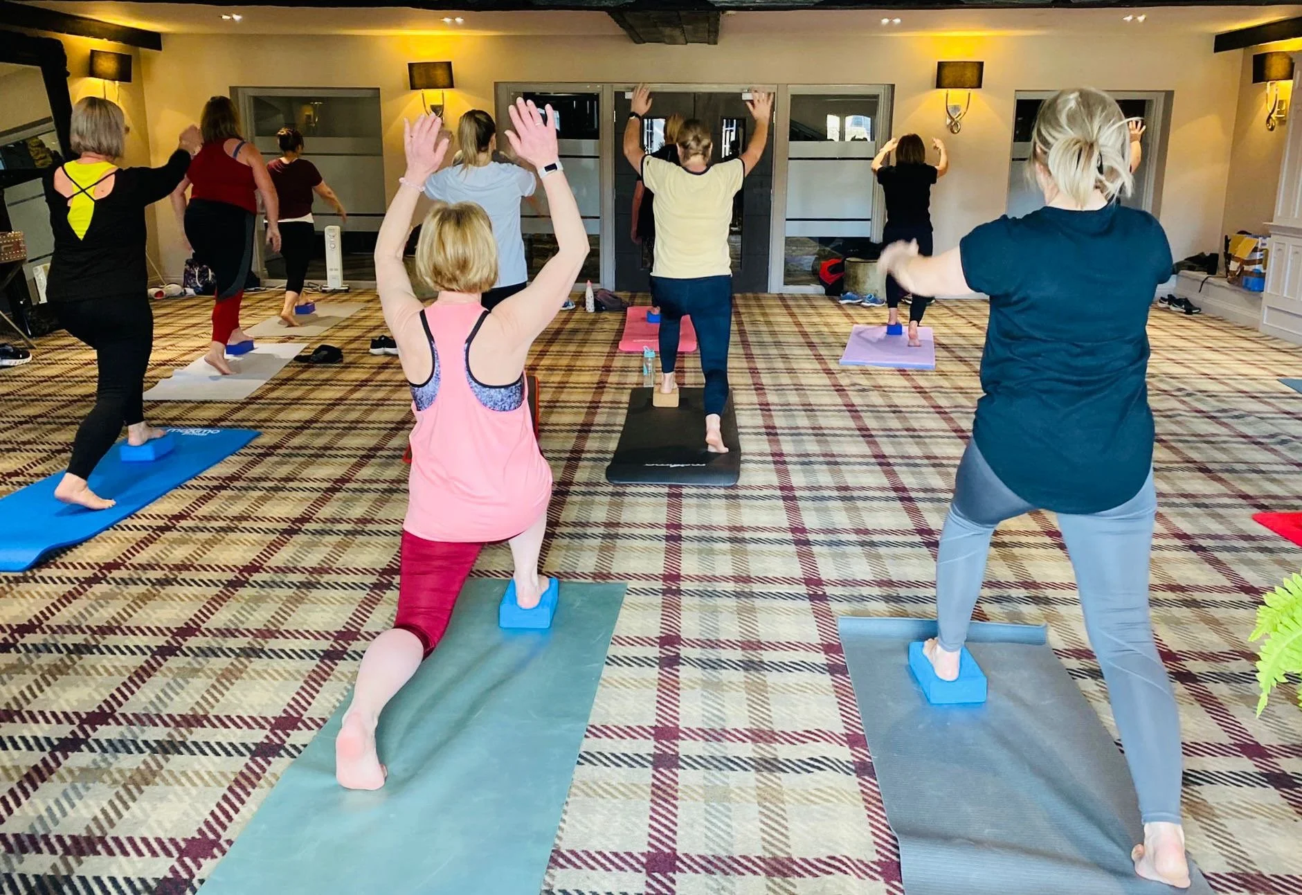 Group of people participating in a yoga class in a spacious, carpeted room, practicing poses on yoga mats with blocks, facing a wall with windows and wall sconces.