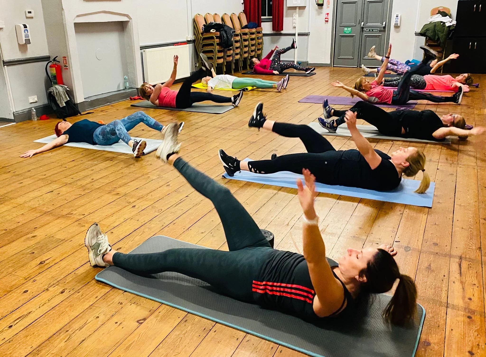 Group of women doing an exercise routine on yoga mats in a room with wooden floors and stacked chairs against the wall.