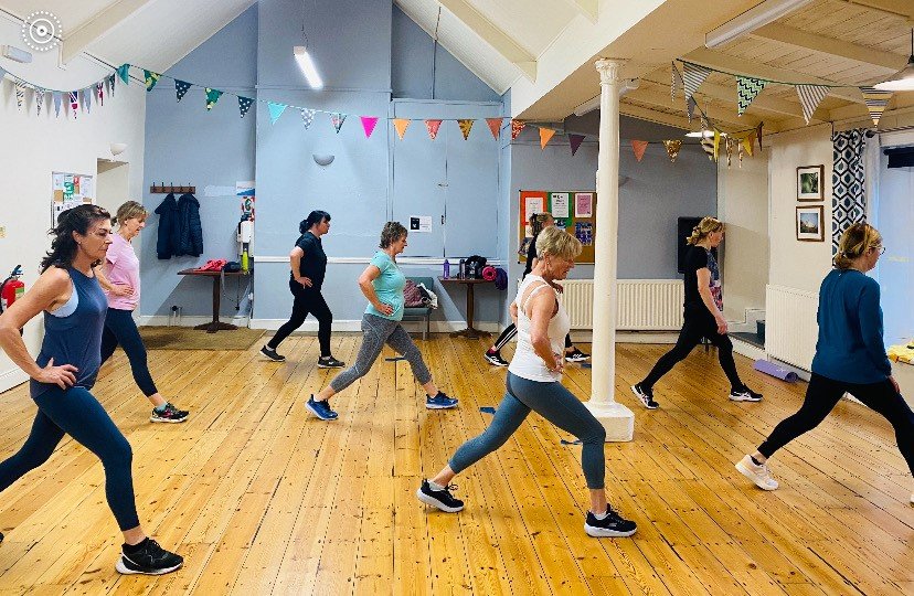 Women participating in a fitness class in a studio with wooden floors, colorful bunting, and framed pictures on the walls.