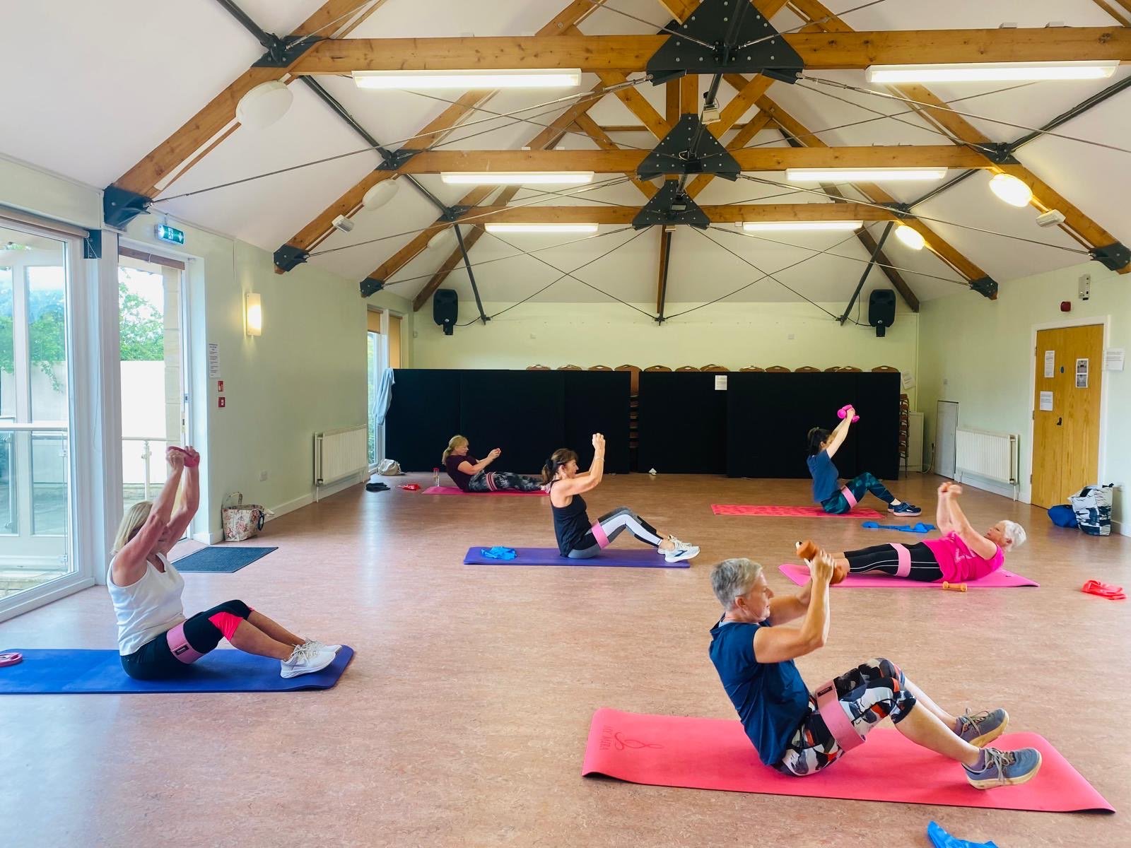 People participating in a seated exercise class with small weights in a spacious, well-lit room with a high wooden ceiling and large windows.
