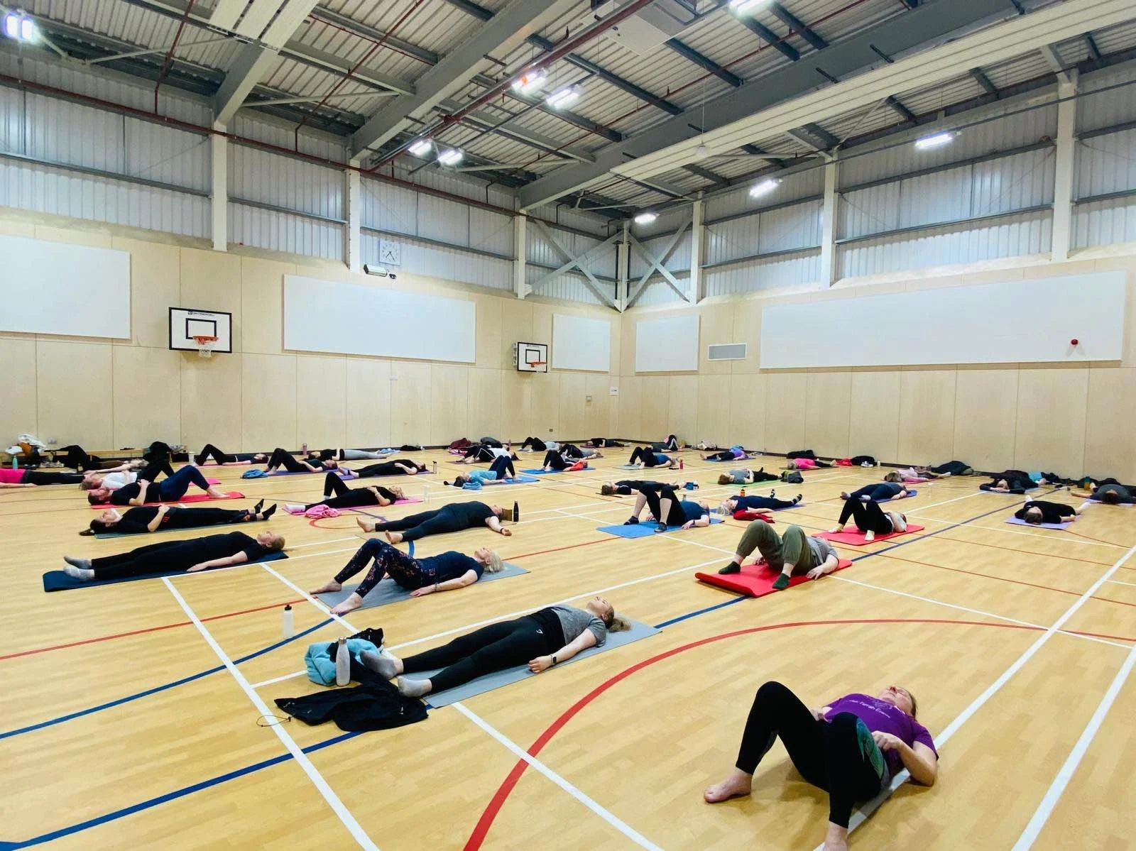 Group of people participating in a yoga or relaxation class in a large indoor gymnasium with wooden floors, basketball hoops, and high ceilings.