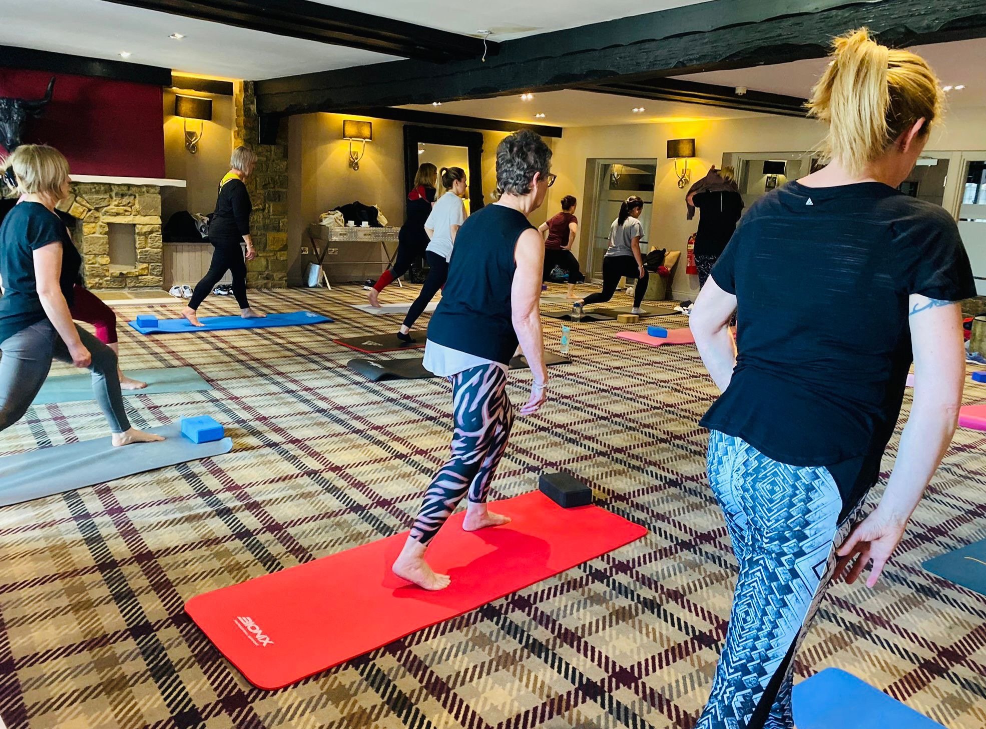 Group of women participating in a yoga class in a room with patterned carpet, some on yoga mats, practicing yoga poses.
