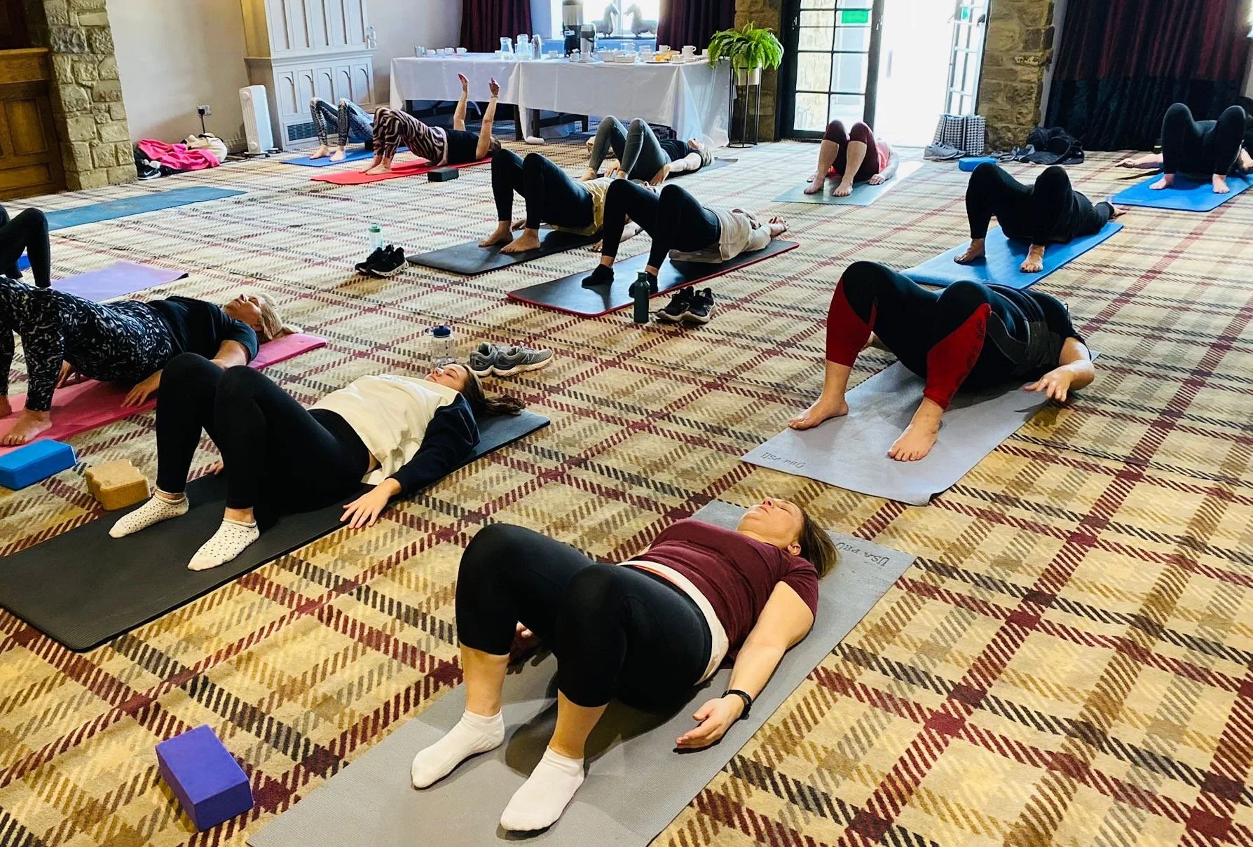 Group of people doing yoga or stretching exercises on mats in a spacious room with checkered carpet, large windows, and a table in the background.