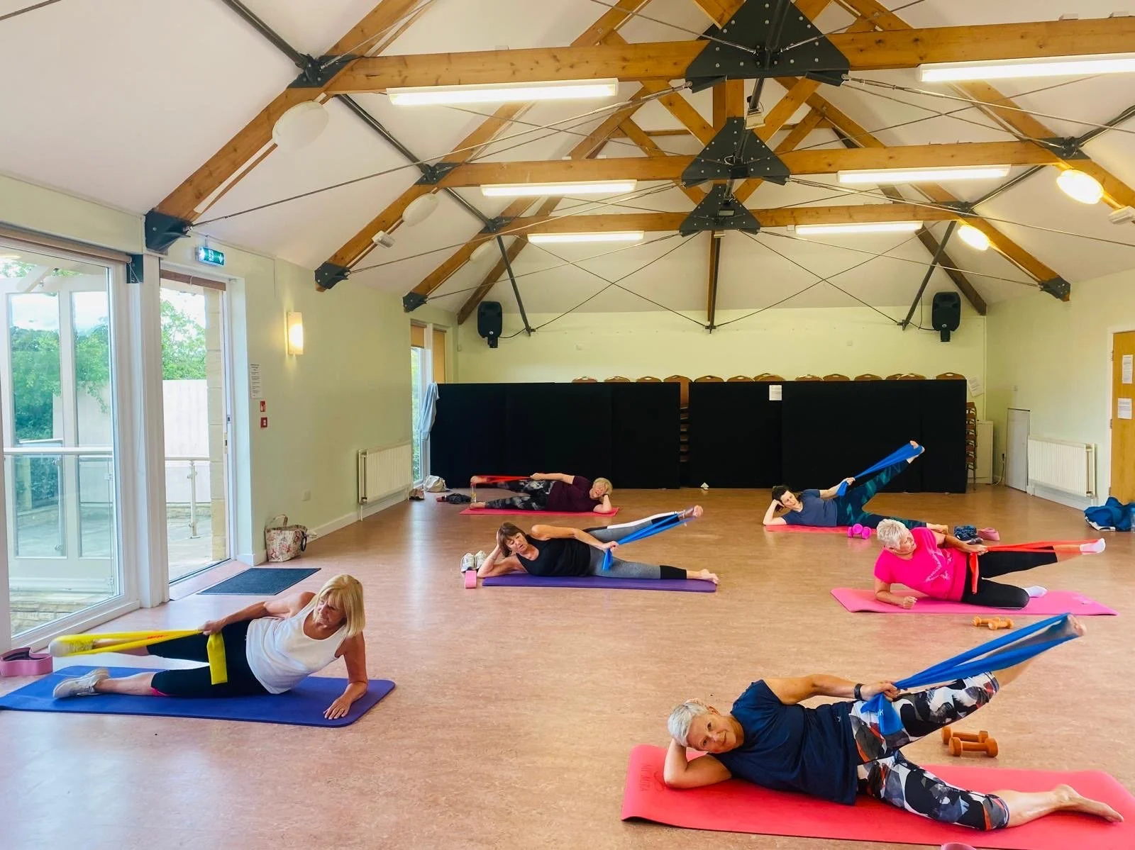 Older women participating in a group yoga or stretching class in a bright, spacious studio with wooden ceiling beams. They are using resistance bands and lying on yoga mats of various colors.