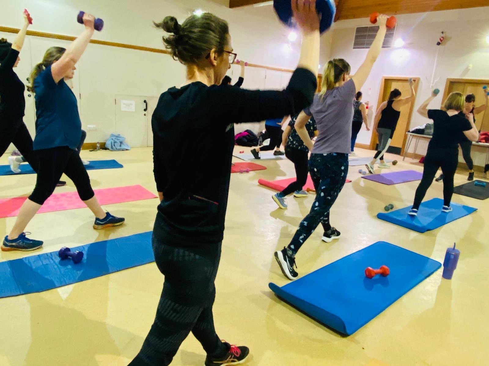 Group of women participating in a fitness class, lifting small dumbbells and using mats in a gym studio.