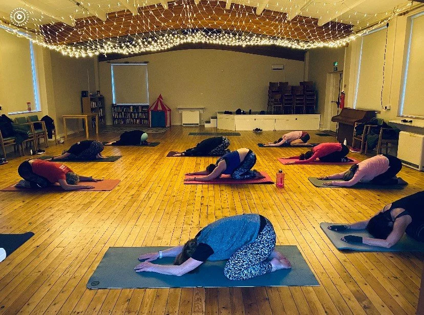 Group of people participating in a yoga class, practicing child’s pose on yoga mats in a spacious, well-lit room decorated with string lights on the ceiling.