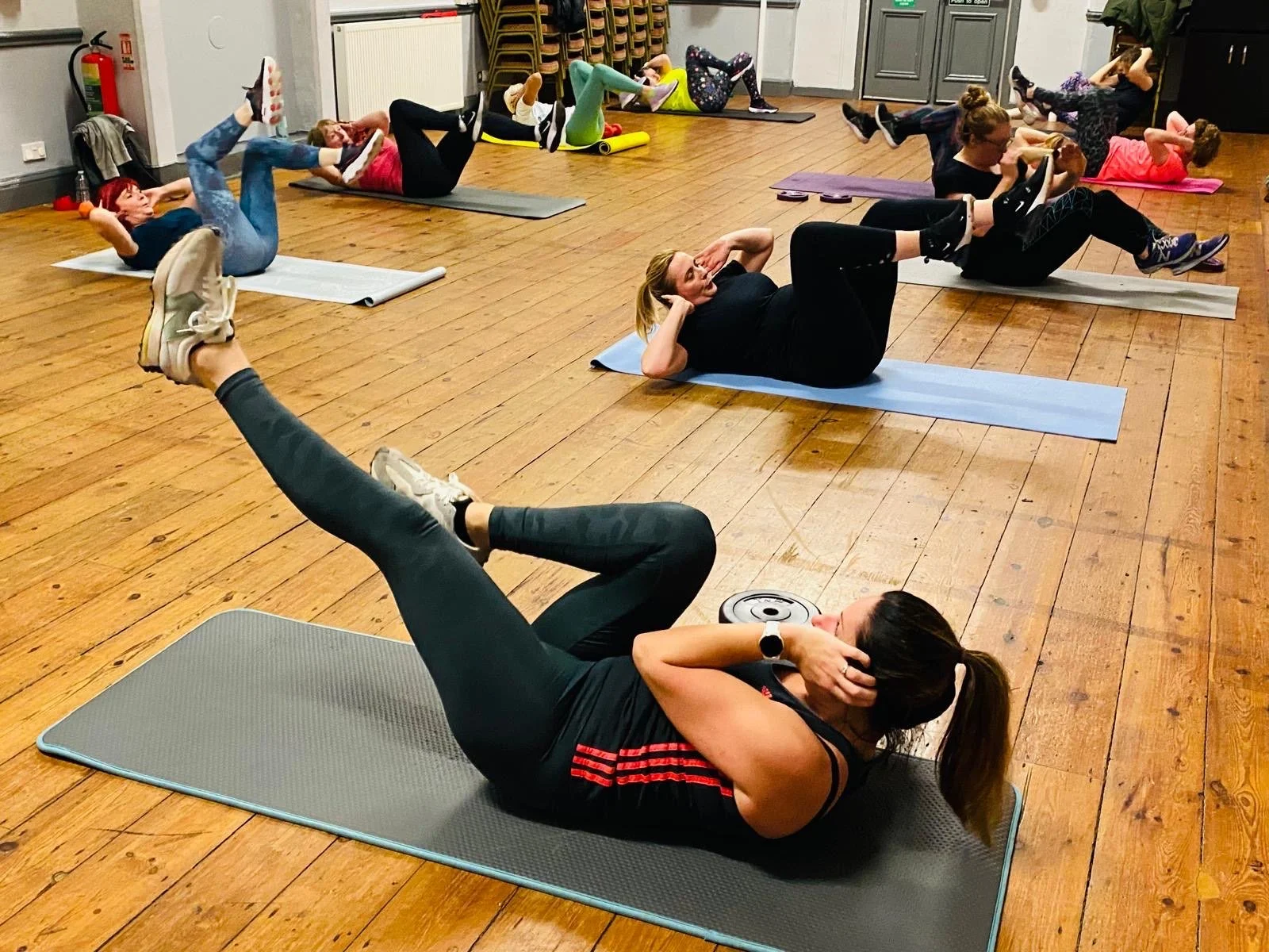 Women participating in a fitness class doing abdominal exercises on mats, with one woman in the foreground using a weight plate as part of her workout.