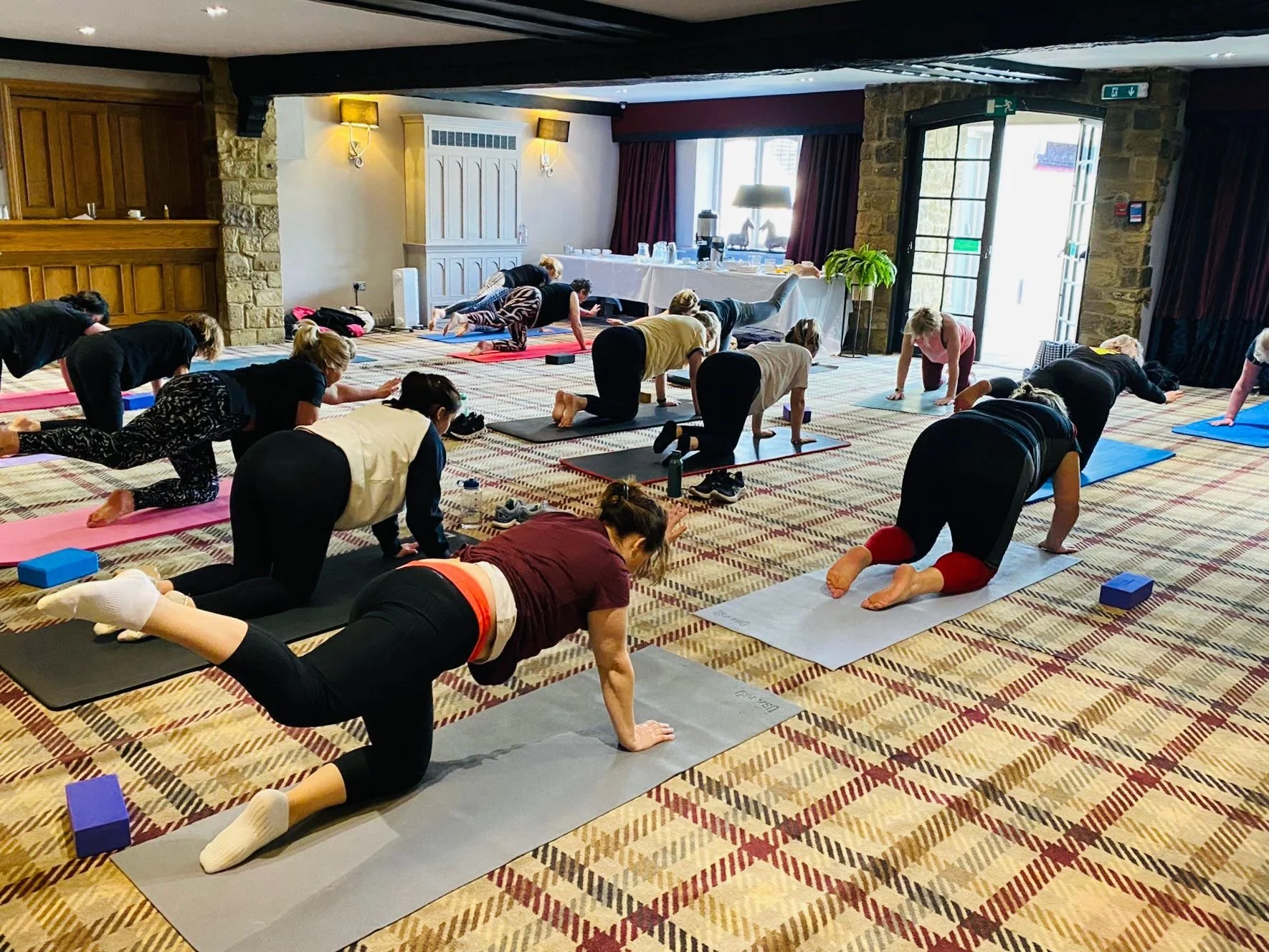 Group of people participating in a yoga class, practicing poses on mats in a spacious room with large windows and a patterned carpet.