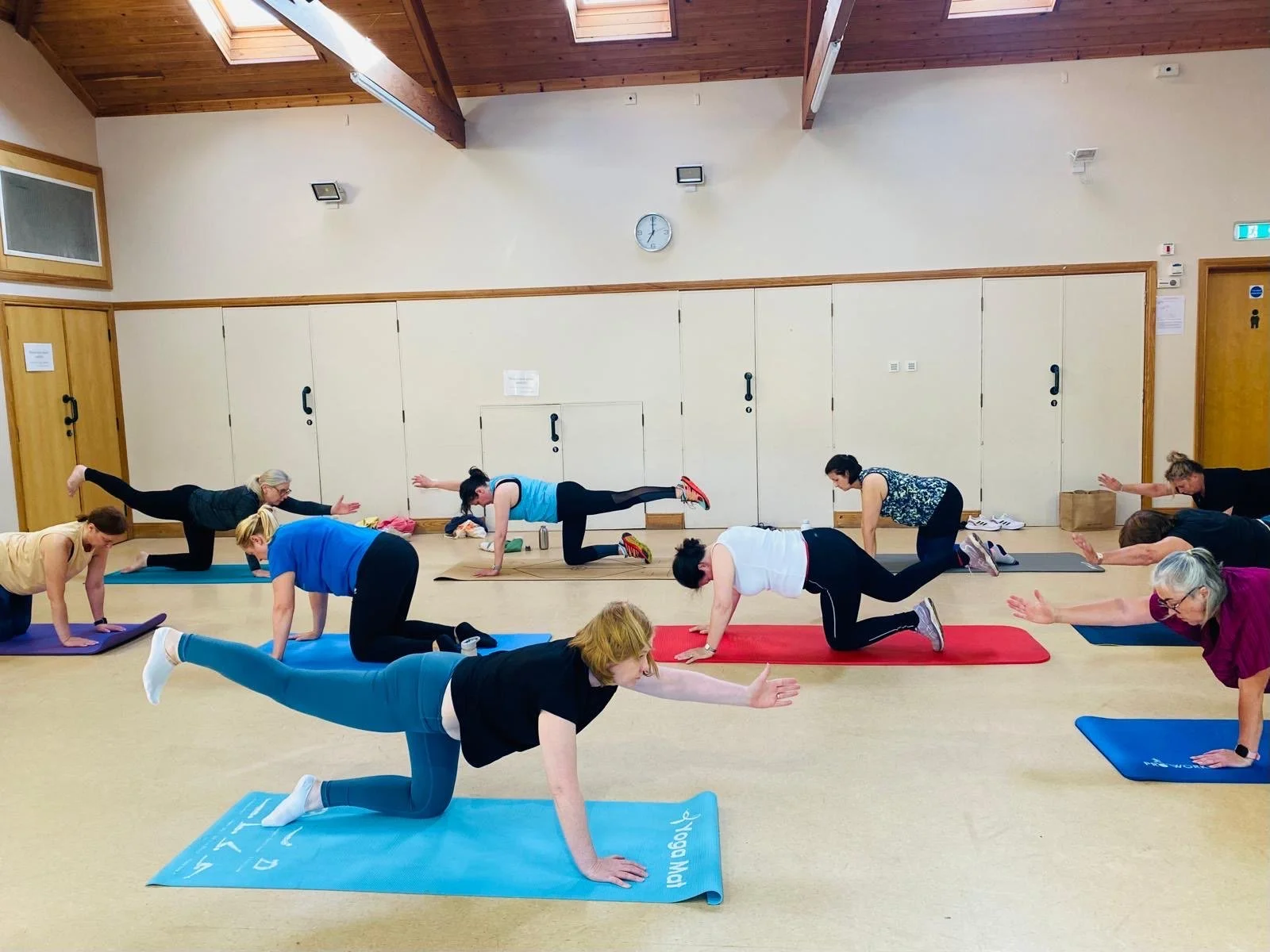 A group of older women participating in a yoga class, performing a pose on their hands and knees on colorful yoga mats in a spacious indoor room with wooden ceiling beams.