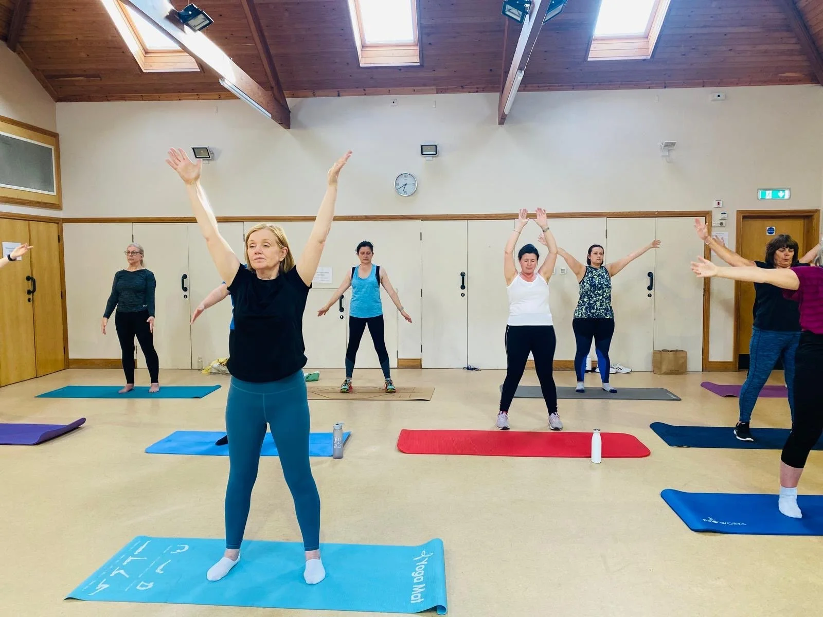 A group of women participating in a yoga class, standing on yoga mats with arms raised or outstretched in a spacious, well-lit room with wooden ceiling beams and skylights.