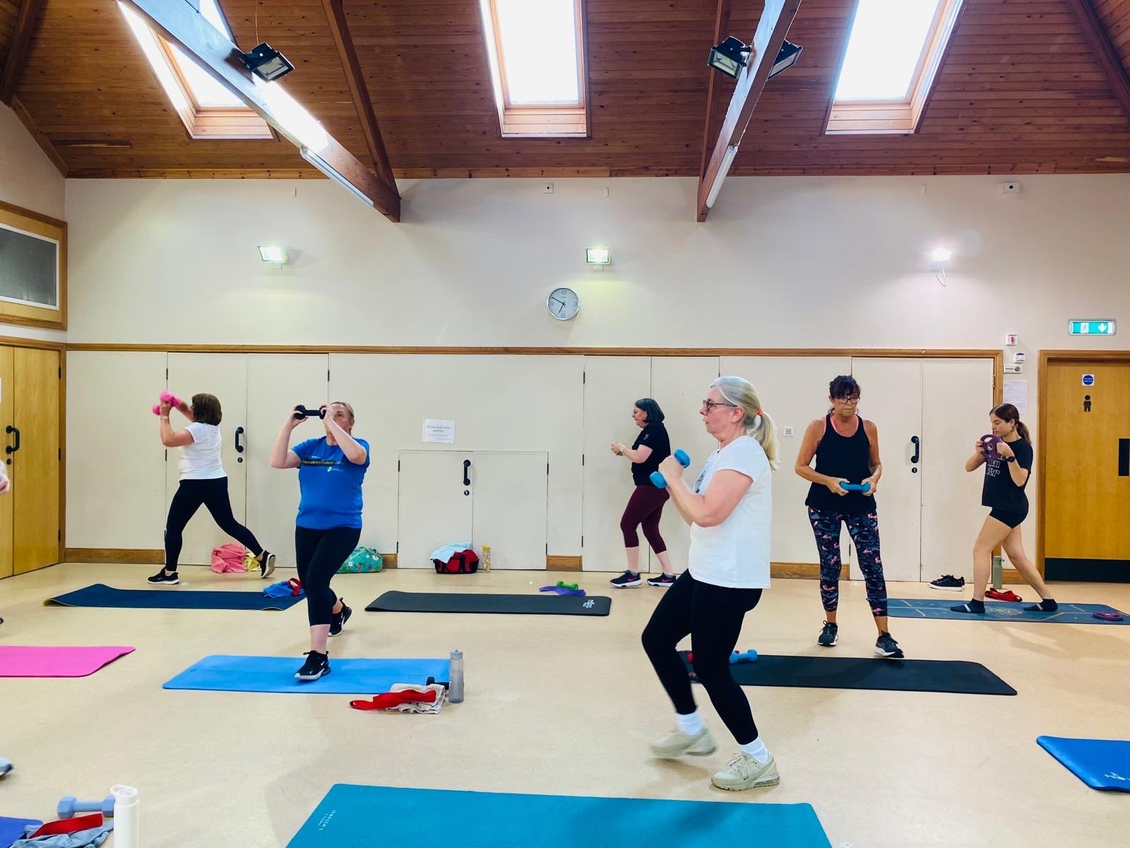 Group of women participating in a fitness class, lifting small dumbbells and using exercise mats in a well-lit indoor studio with wooden ceiling and skylight windows.