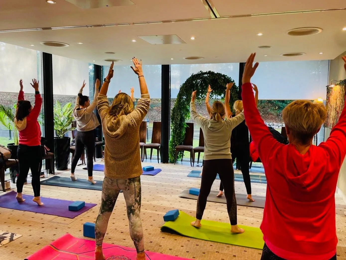 Group of people practicing yoga in a bright indoor space with large windows, greenery outside, and yoga mats.