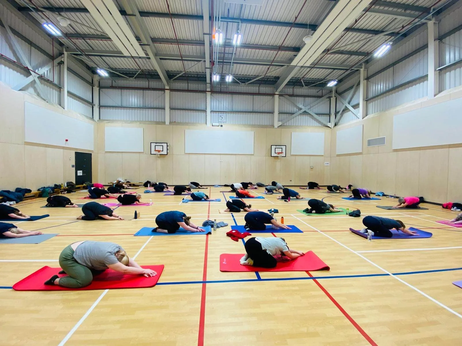 Group of people practicing yoga in a gymnasium, doing child's pose on yoga mats arranged in rows on a light wood floor, with basketball hoops and a clock on the wall.