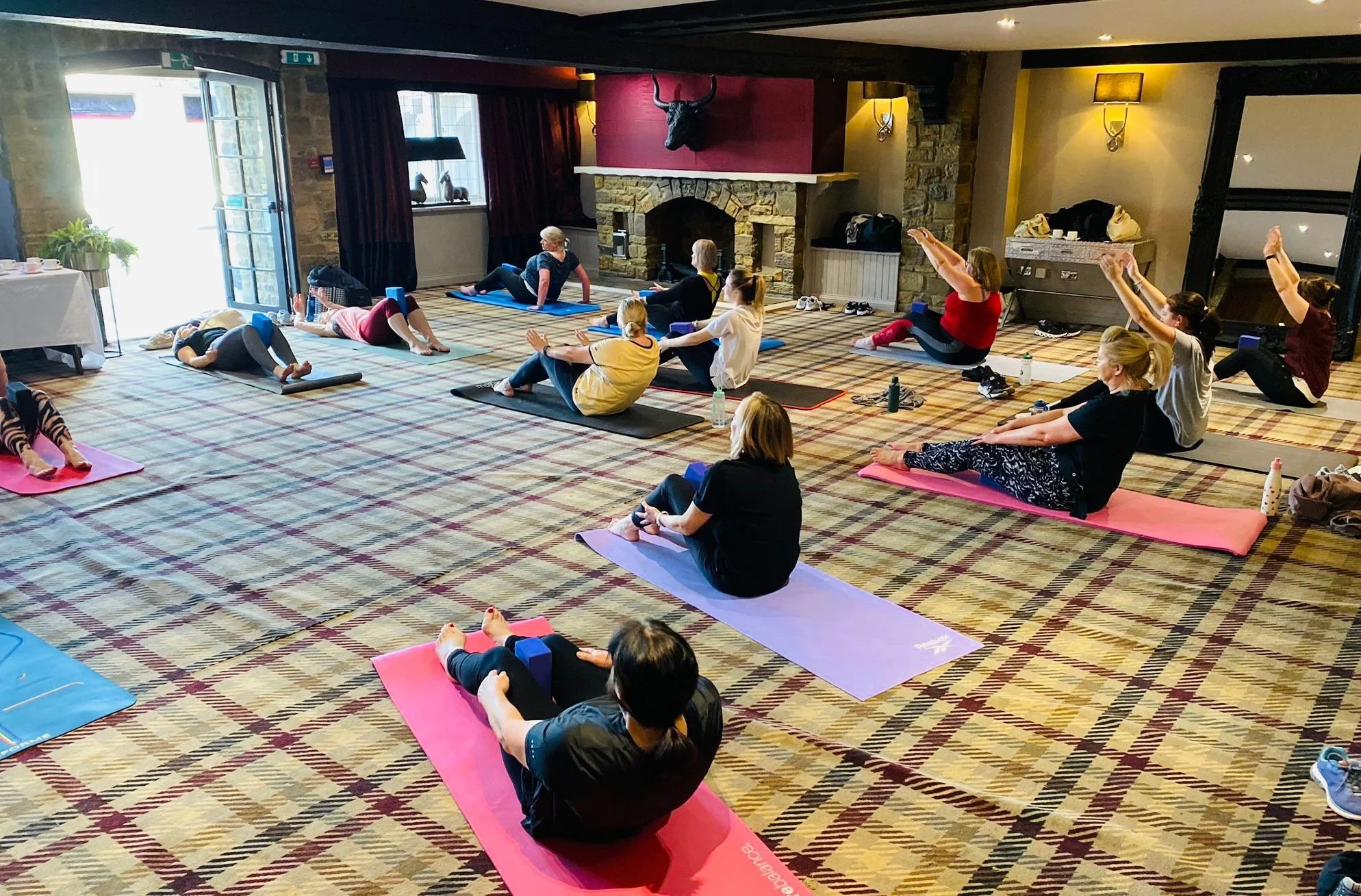 Interior of a yoga or exercise class in a rustic room with stone fireplace, carpeted floor, and people on yoga mats stretching and doing poses.