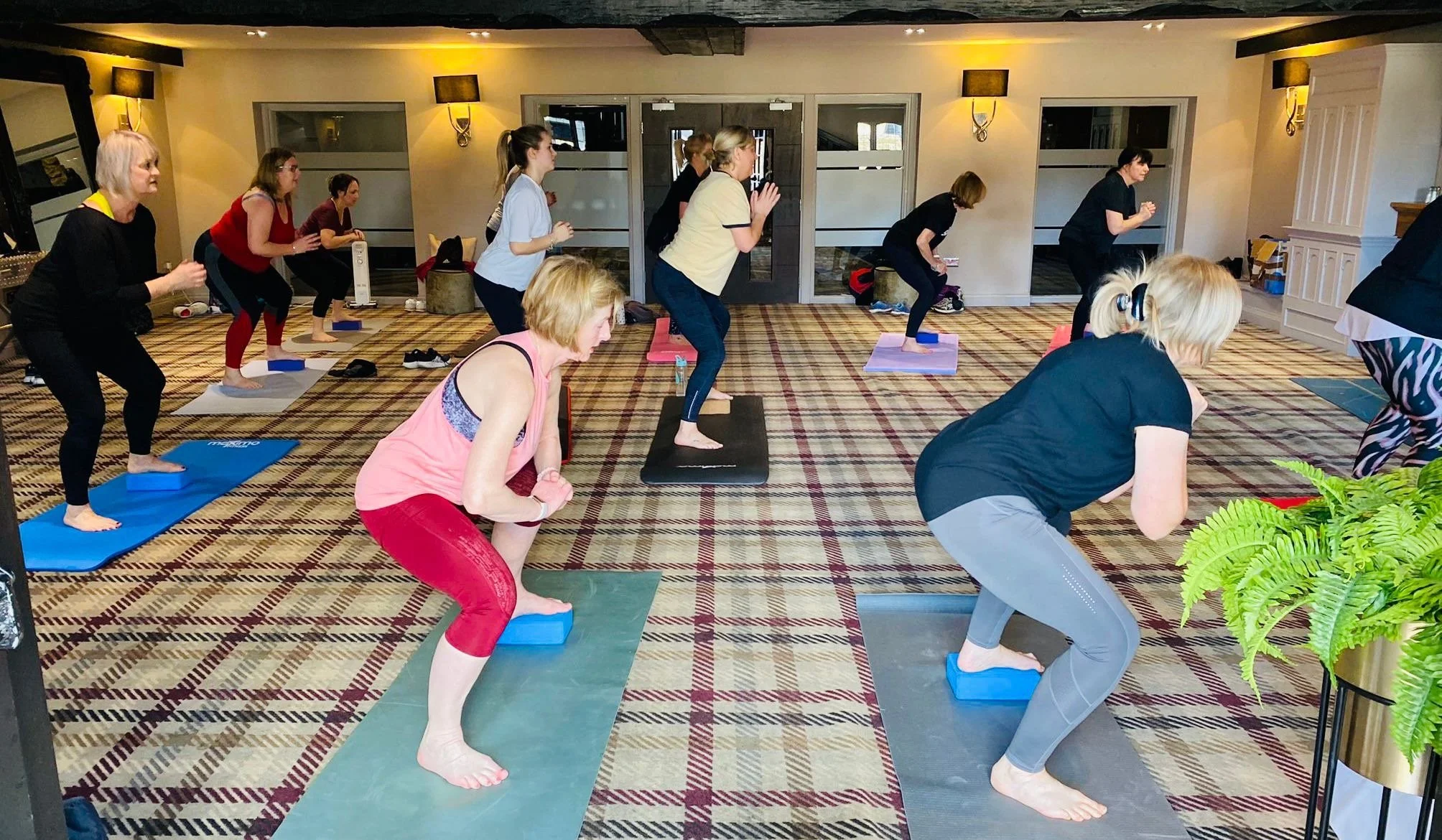 Group of women participating in a yoga or exercise class in a room with yellow walls, wall-mounted lamps, and a checkered carpet.