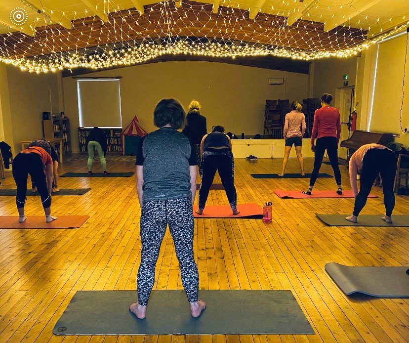 People participating in a yoga class, standing on yoga mats in a well-lit room with decorated ceiling lights.