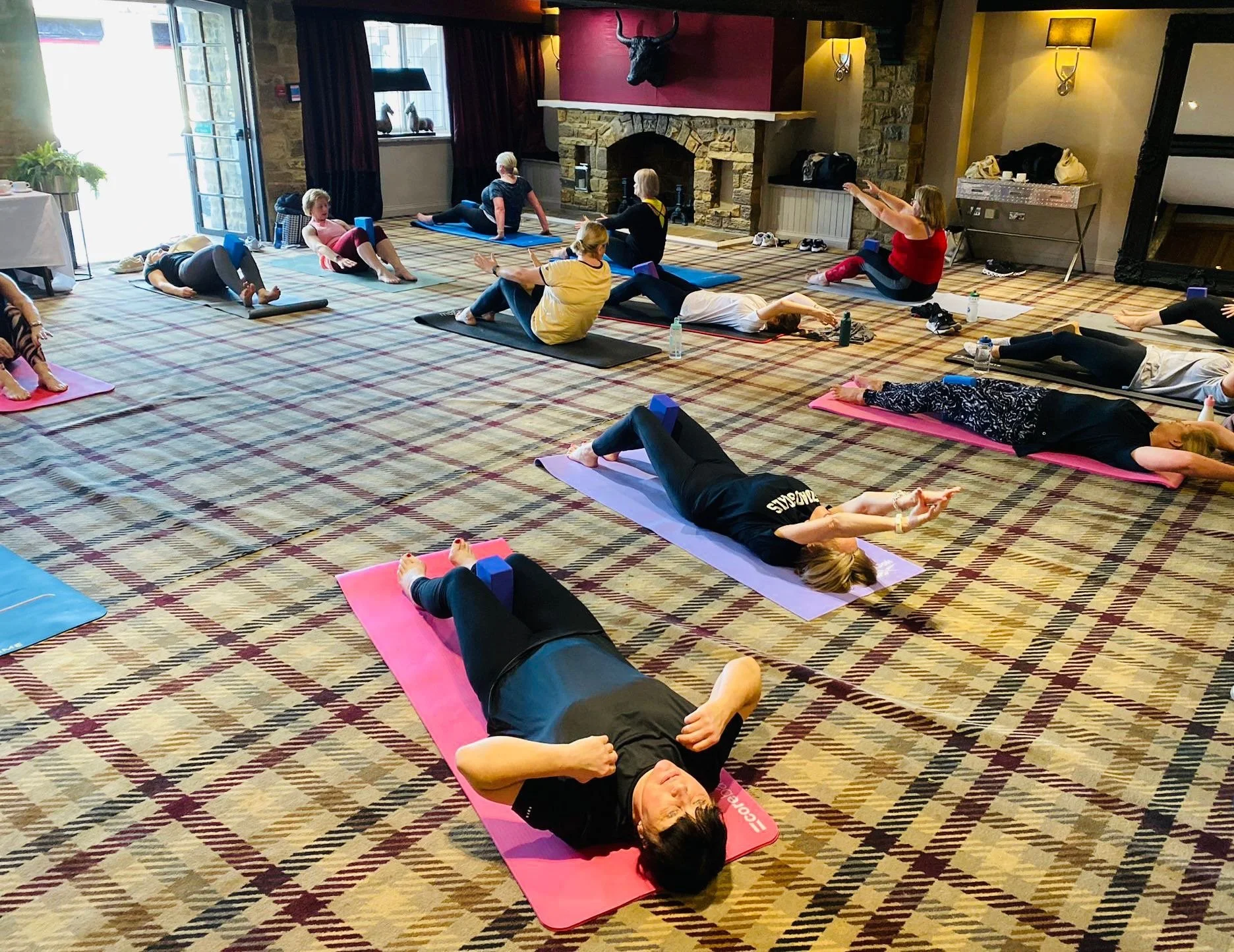 A group of people participating in a yoga class in a spacious indoor room with large windows, a fireplace, and a plaid carpet. They are lying on yoga mats in a relaxation pose, with several instructors leading the session.