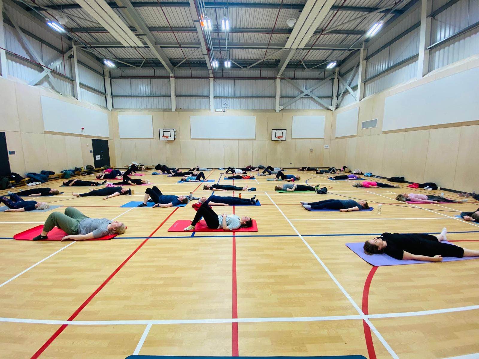 People participating in a yoga or stretching class in a gymnasium with wooden floors, high ceilings, and two basketball hoops.