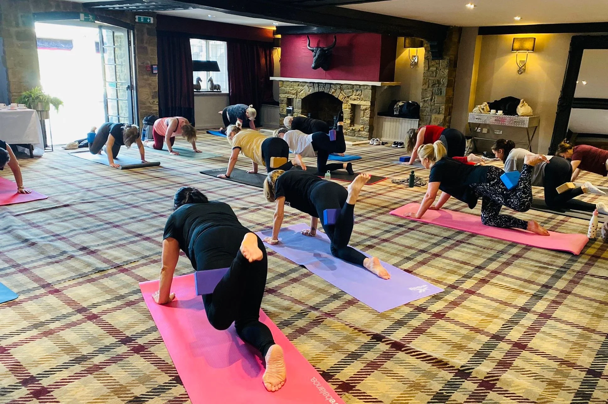 People participating in a yoga or Pilates class in a spacious room with a stone fireplace, a mounted animal head, large windows, and colorful mats.