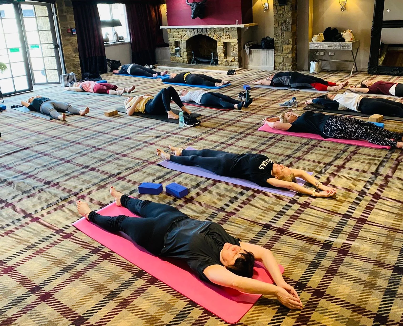 Group of people participating in a yoga or meditation class, lying on yoga mats on a patterned carpeted floor in a spacious room with stone walls and large windows.