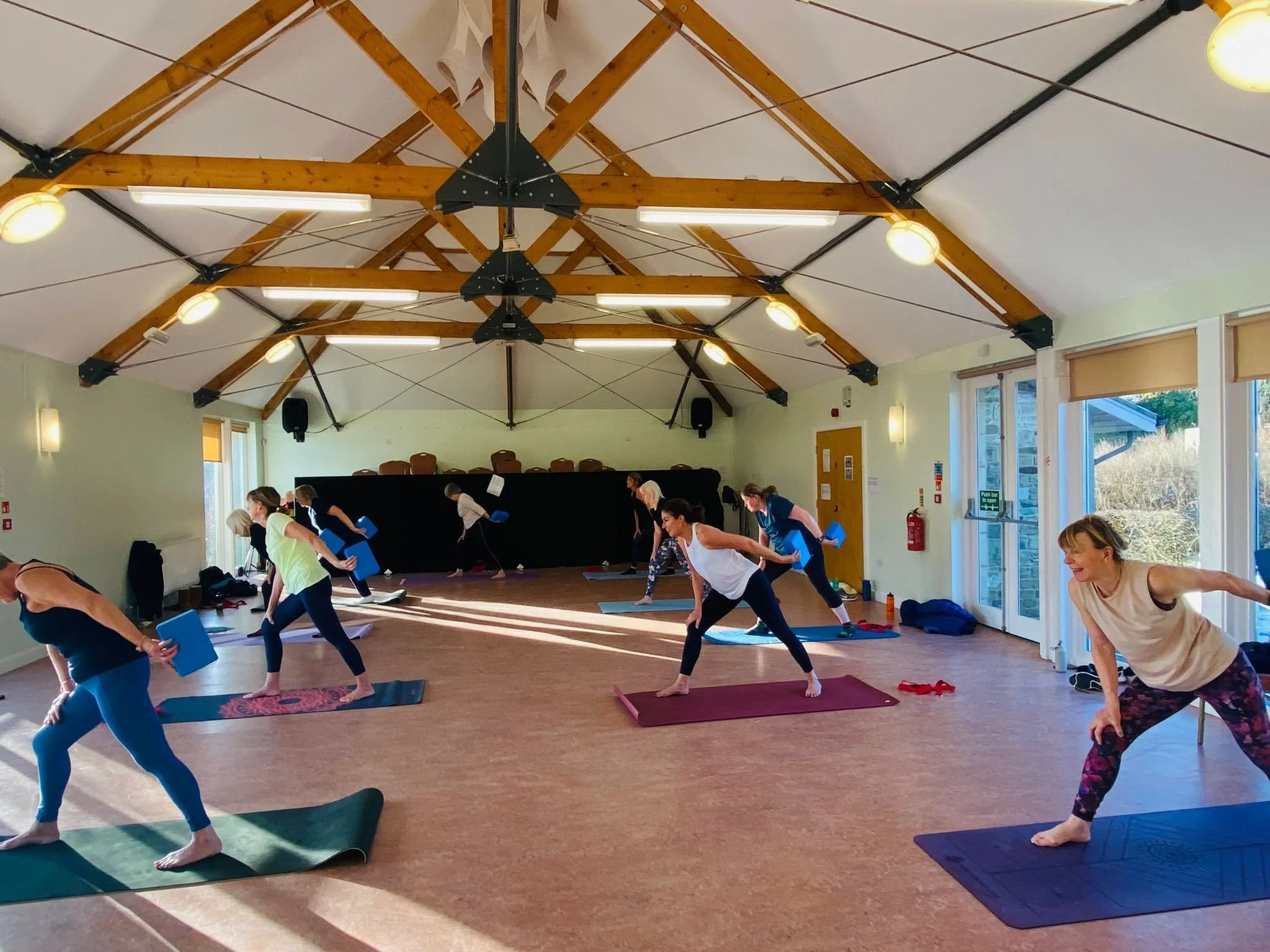A group of women participating in a yoga or fitness class in a spacious room with wooden beams on the ceiling and large windows.