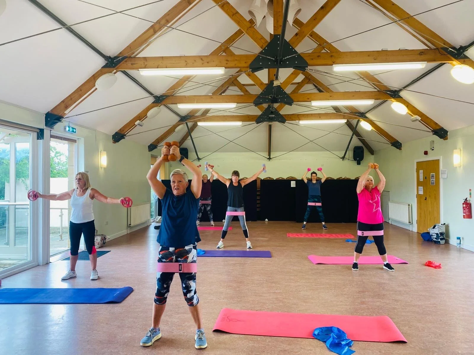 Group of older women participating in a fitness class with dumbbells and resistance bands in a spacious room with wooden ceiling beams.
