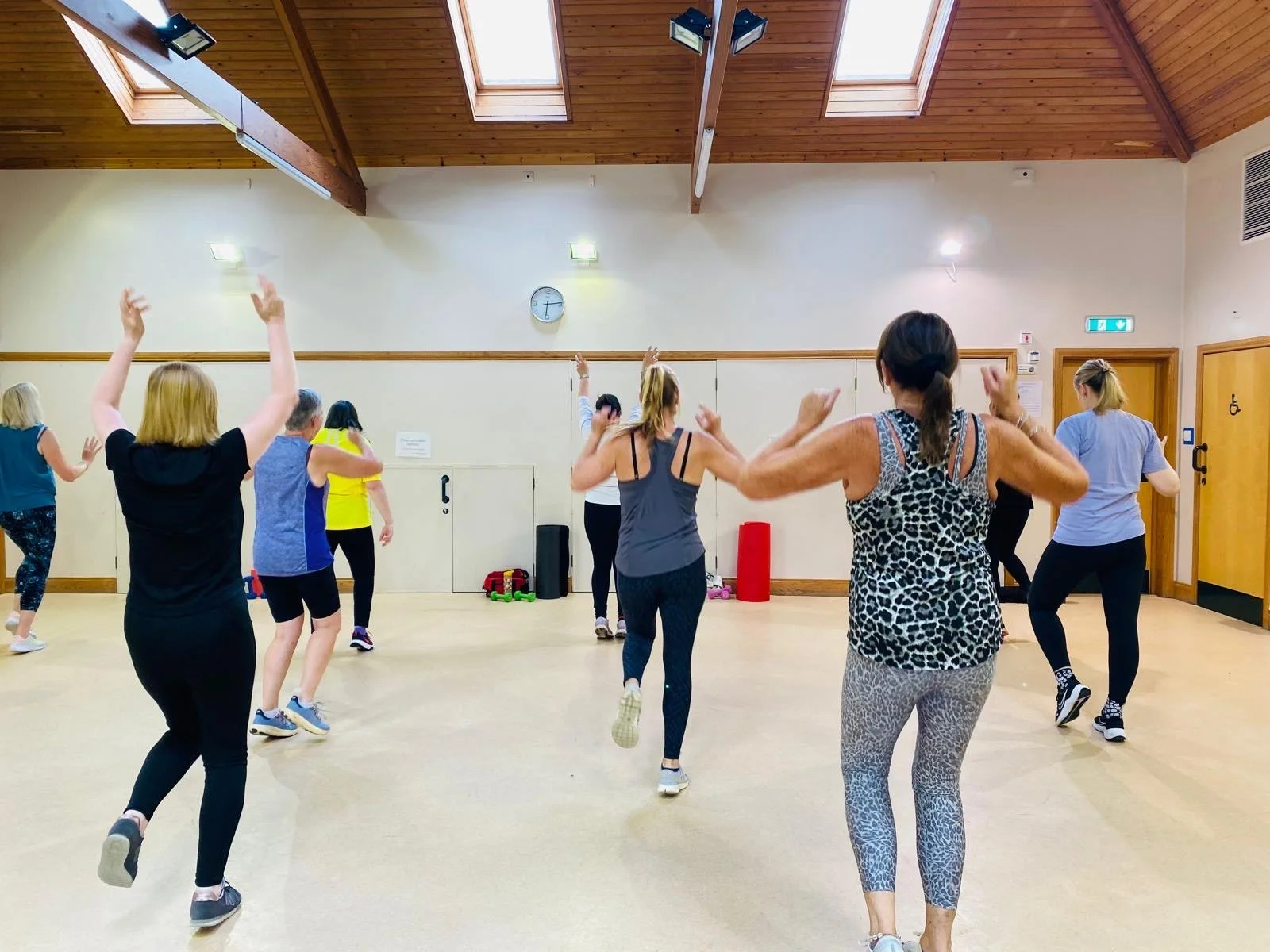 Group of people participating in a dance or exercise class in a spacious, well-lit room with wooden ceiling and light-colored walls.