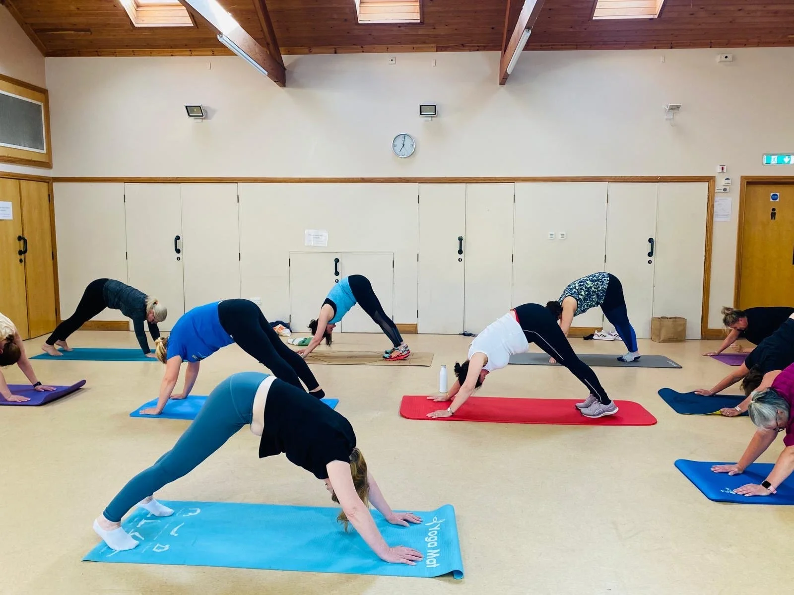People participating in a yoga class in a spacious room with wooden ceiling beams and beige walls, all in downward-facing dog pose on colorful mats.