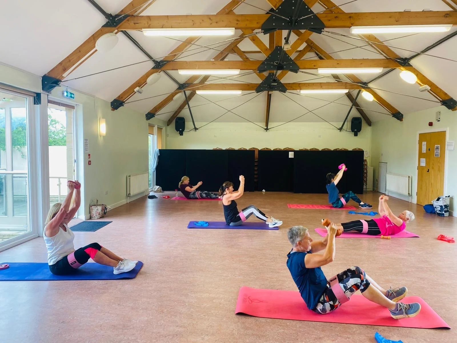 Older adults participating in a seated exercise class in a spacious wooden-ceiling room with windows, lifting pink and orange weights on exercise mats.