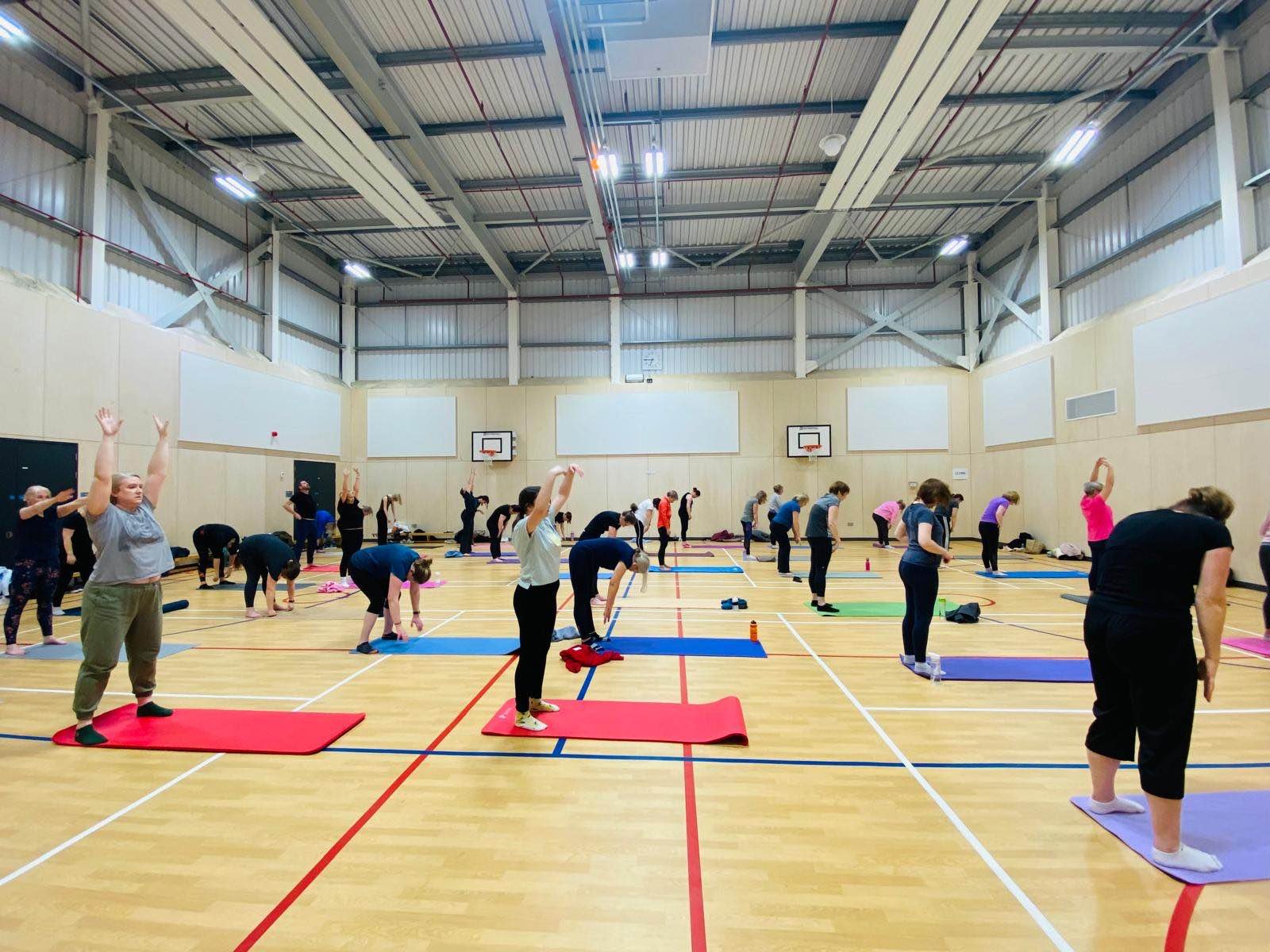People participating in a yoga class inside a gymnasium with wooden walls and a high ceiling, all on yoga mats.