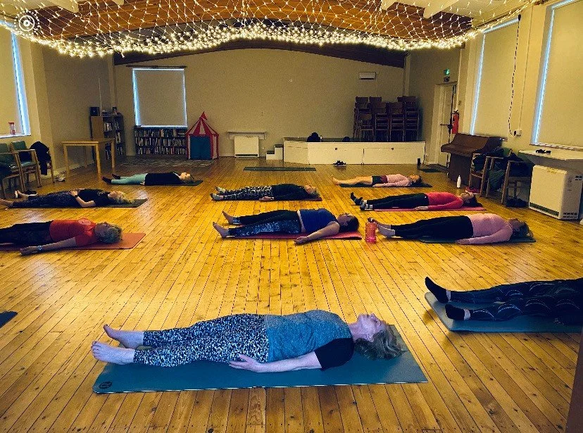 People participating in a yoga or meditation class lying on yoga mats in a spacious room with wooden floors, draped with string lights on the ceiling.