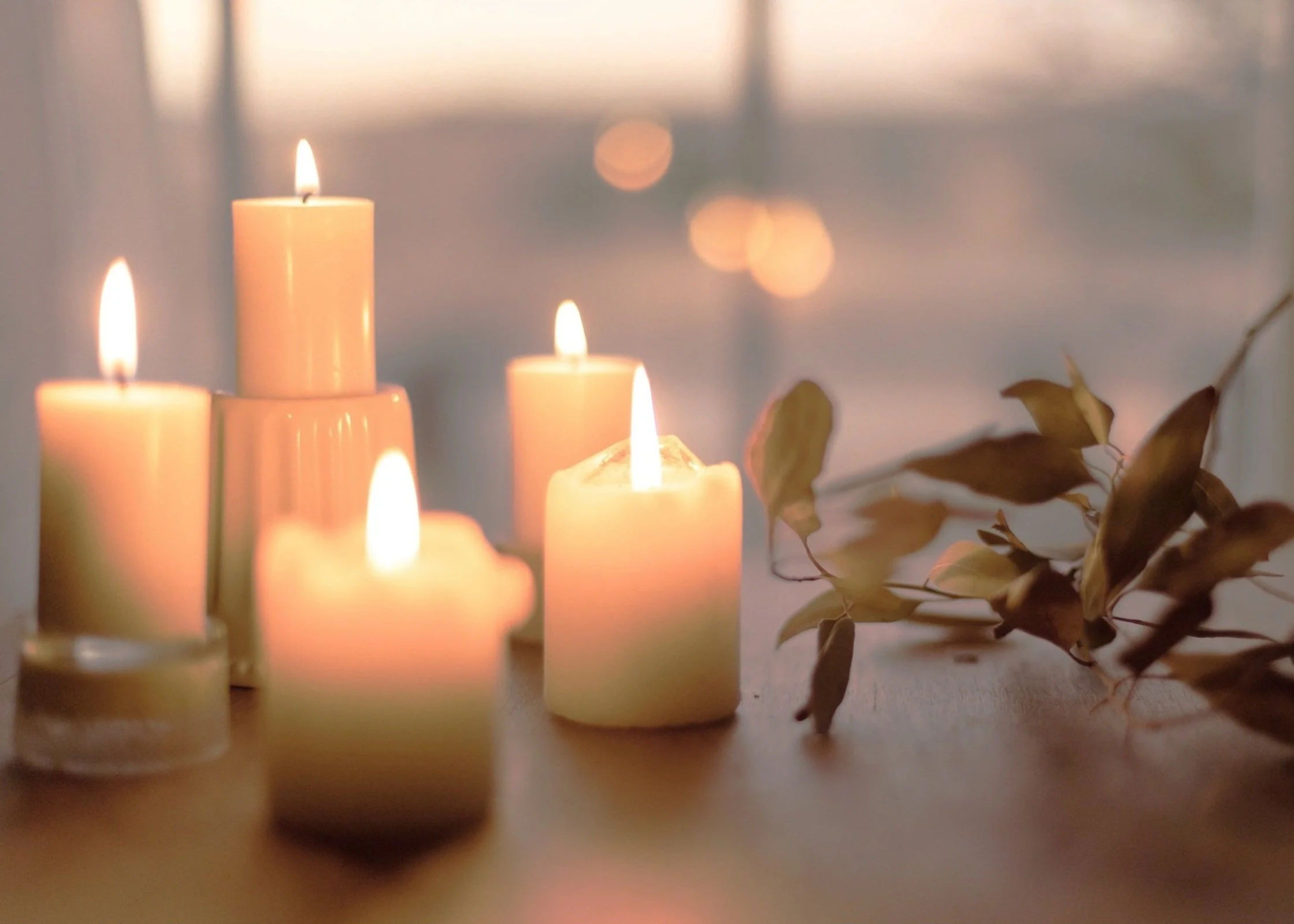 Several lit candles and dried branches on a surface, with a blurred background of warm lights.