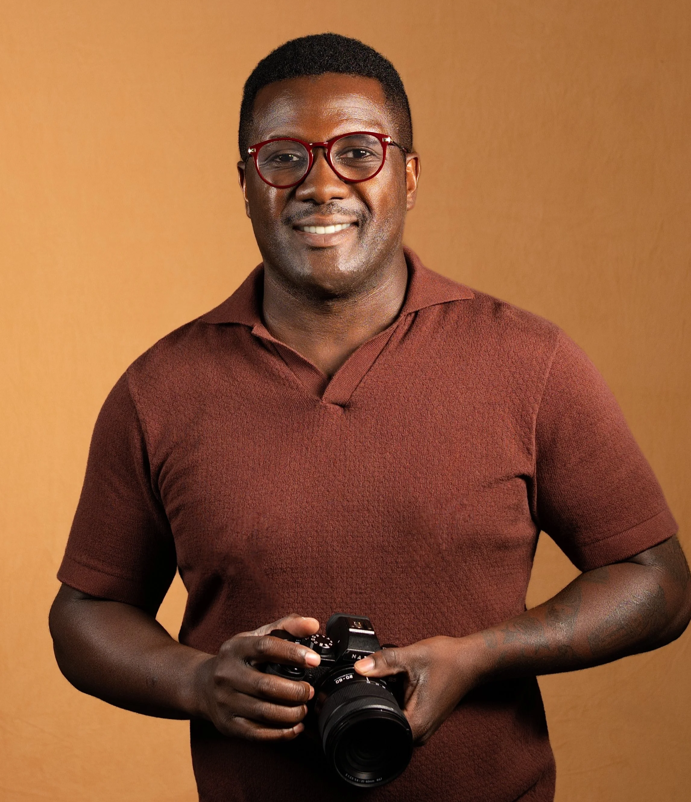 A Black man with short hair wearing red glasses and a maroon shirt, holding a camera, standing against an orange-brown background.