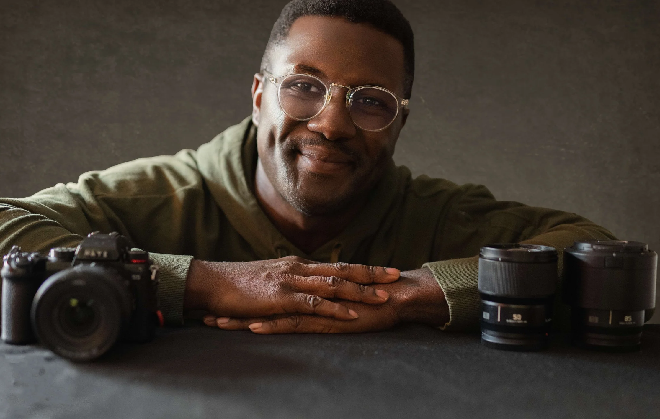 A young man wearing glasses and a green hoodie resting his folded arms on a black surface, with camera equipment and lenses in front of him, smiling softly.